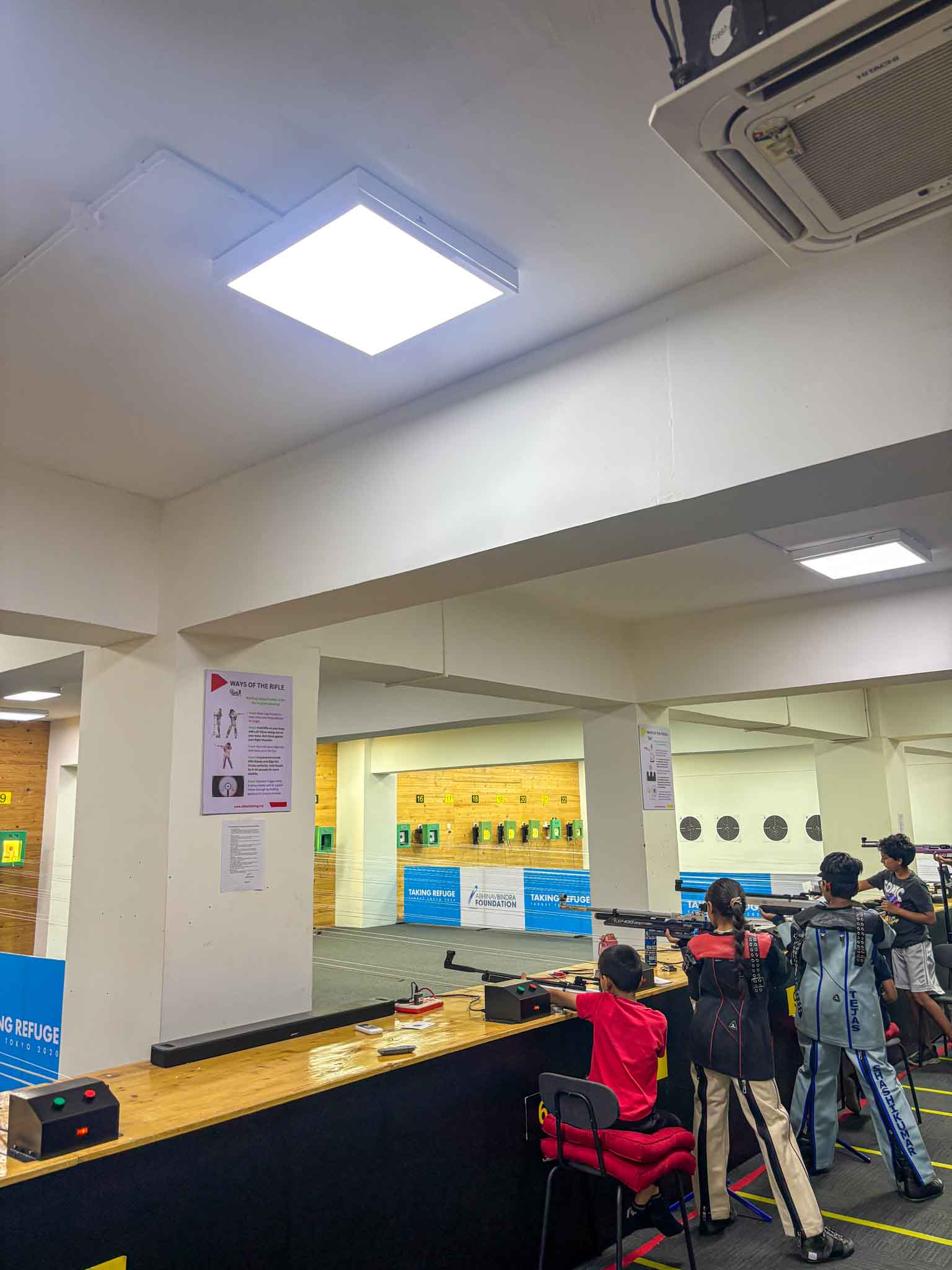 Children practicing rifle shooting at a well-lit indoor shooting range in Bengaluru, wearing shooting gear.